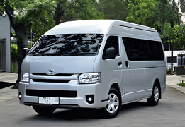Toyota Hiace High Grade warna putih difoto dari sudut depan samping, menampilkan bodi van panjang dengan kapasitas besar, kaca jendela lebar di setiap baris kursi, dan desain grille depan yang tegas. Sebagai armada sewa mobil di Medan, Hiace menjadi pilihan utama untuk rombongan besar seperti study tour, rombongan wisata ke Danau Toba/Berastagi, antar-jemput bandara Kualanamu, hingga acara pernikahan dan perusahaan yang membutuhkan kapasitas hingga 15 penumpang