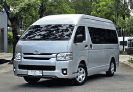 Toyota Hiace High Grade warna putih difoto dari sudut depan samping, menampilkan bodi van panjang dengan kapasitas besar, kaca jendela lebar di setiap baris kursi, dan desain grille depan yang tegas. Sebagai armada sewa mobil di Medan, Hiace menjadi pilihan utama untuk rombongan besar seperti study tour, rombongan wisata ke Danau Toba/Berastagi, antar-jemput bandara Kualanamu, hingga acara pernikahan dan perusahaan yang membutuhkan kapasitas hingga 15 penumpang